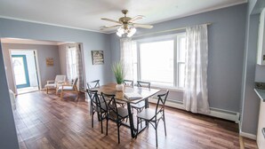 Un espace repas comprenant une table et des chaises en bois, un ventilateur de plafond et de grandes fenêtres avec des rideaux.