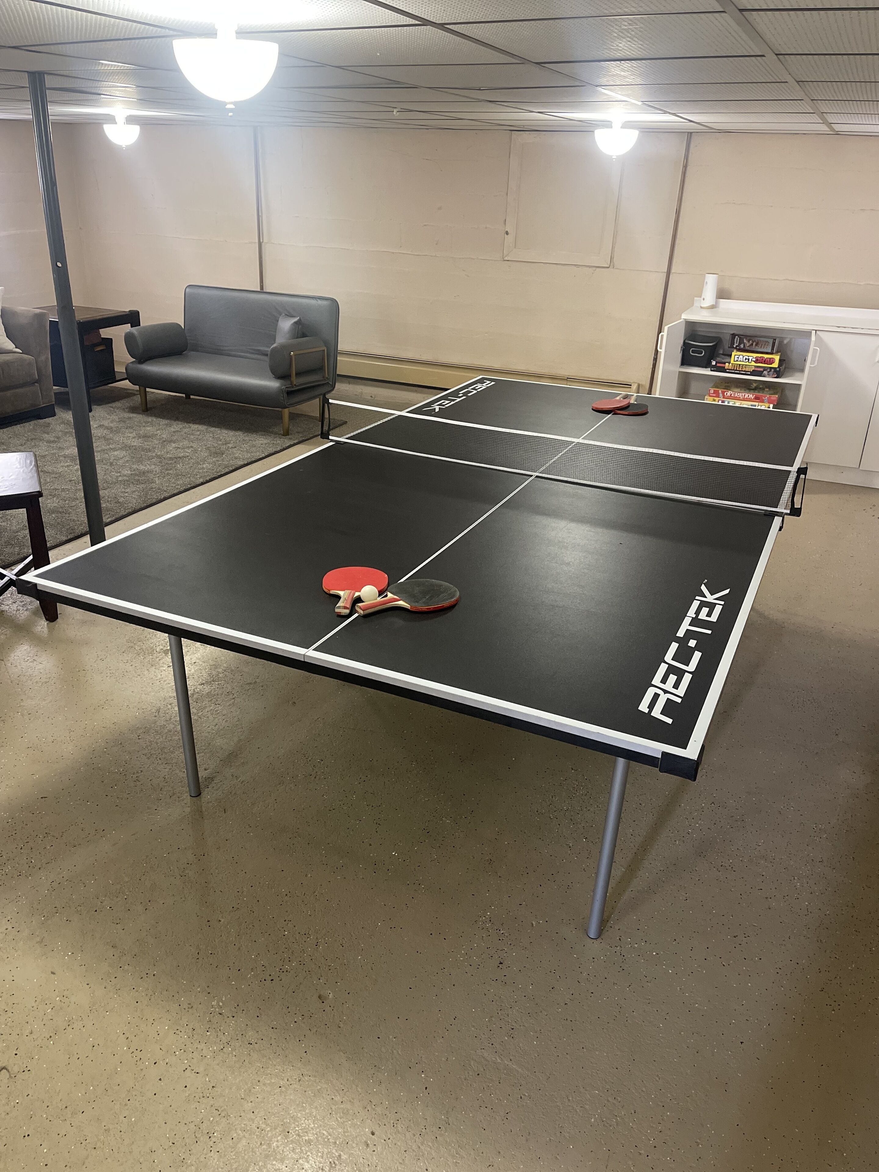 A ping pong table with a net, a red paddle, and a ball. There are two chairs and a small table in the background.