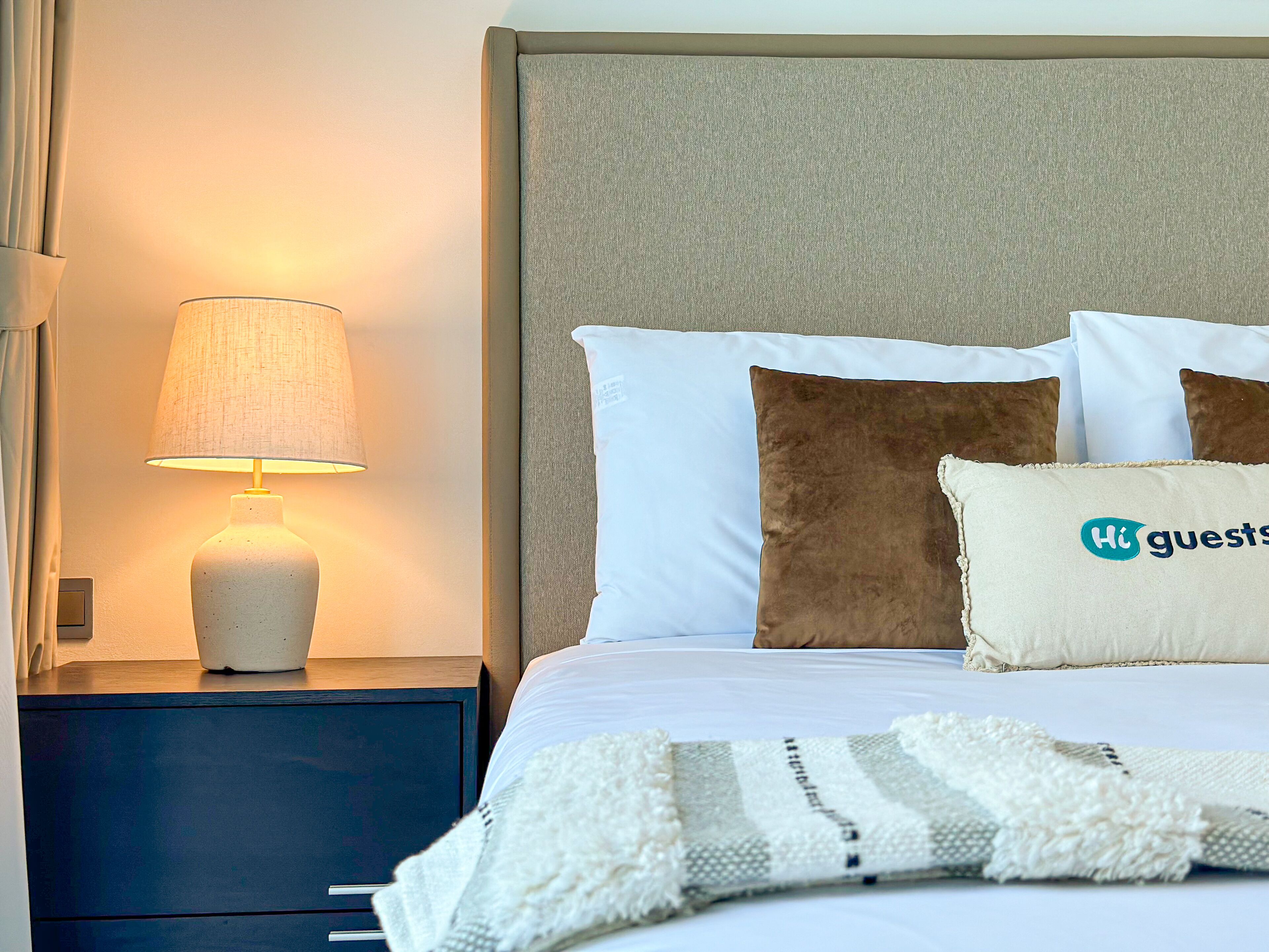 A neatly made bed with white and brown pillows, a white throw blanket, and a lit table lamp on a bedside table.