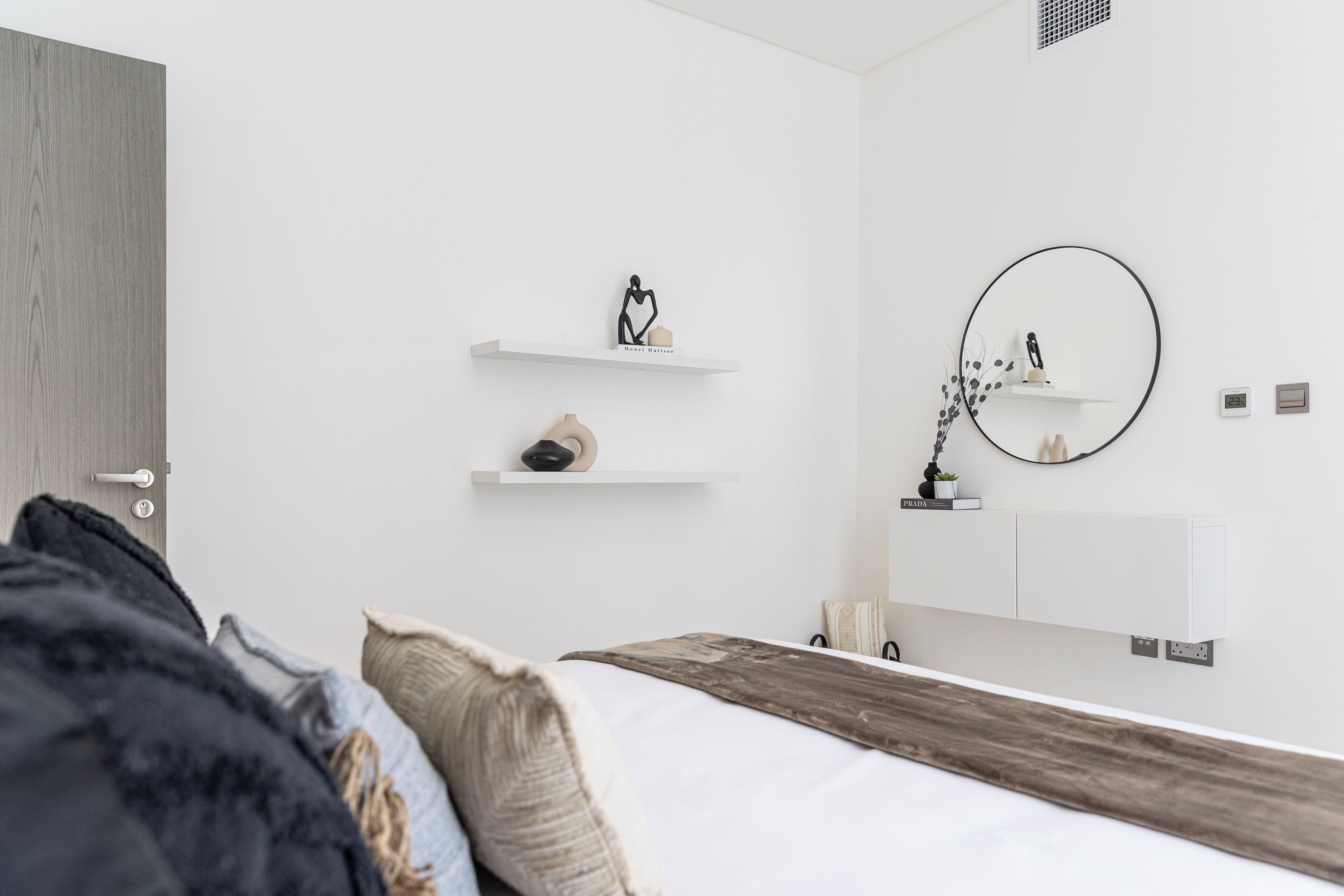 A modern bedroom with a wooden headboard, white walls, and minimalist decor including a round mirror and two floating shelves.