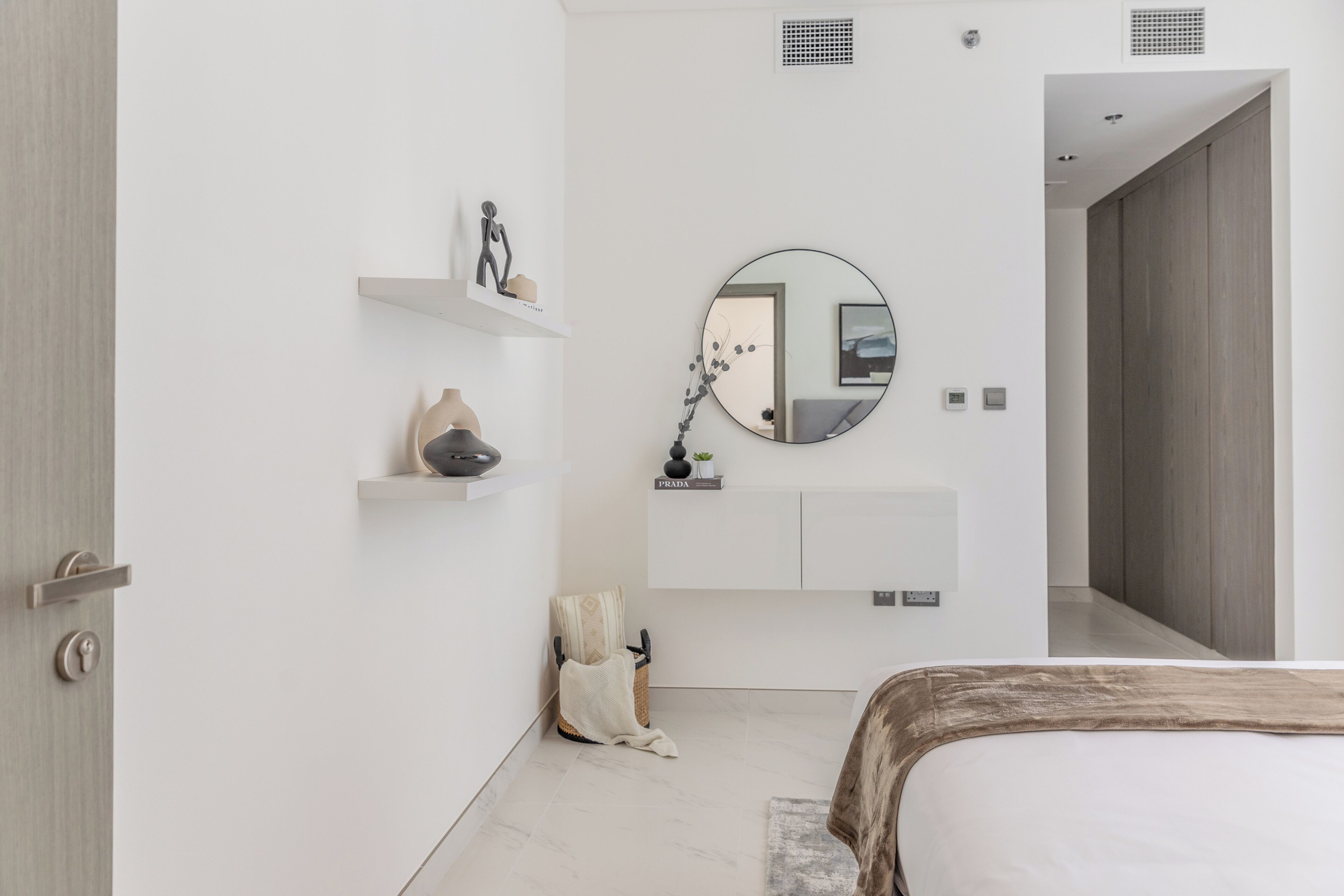 A modern bedroom with a bed, a round mirror, a shelf with decorative items, and a door.