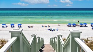 Ein Strand mit einer Holztreppe, die zum Sand führt, umgeben von blauen Sonnenschirmen und Liegestühlen.