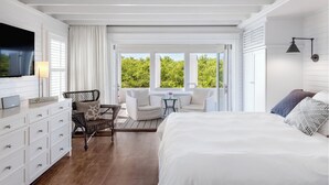 Ein Schlafzimmer mit einem großen Bett, einem Fernseher, einem Schreibtisch mit zwei Stühlen und Blick auf Grün hinter den Fenstern.