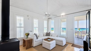 Ein geräumiges Wohnzimmer mit einem weißen Modulsessel, einem schwarzen Kaminofen und großen Fenstern mit Blick auf den Strand.