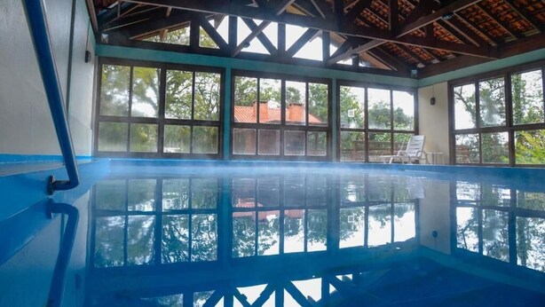 An indoor swimming pool with a view of trees and a building through large windows. - Residencial Pousada do Serrano (Gramado)