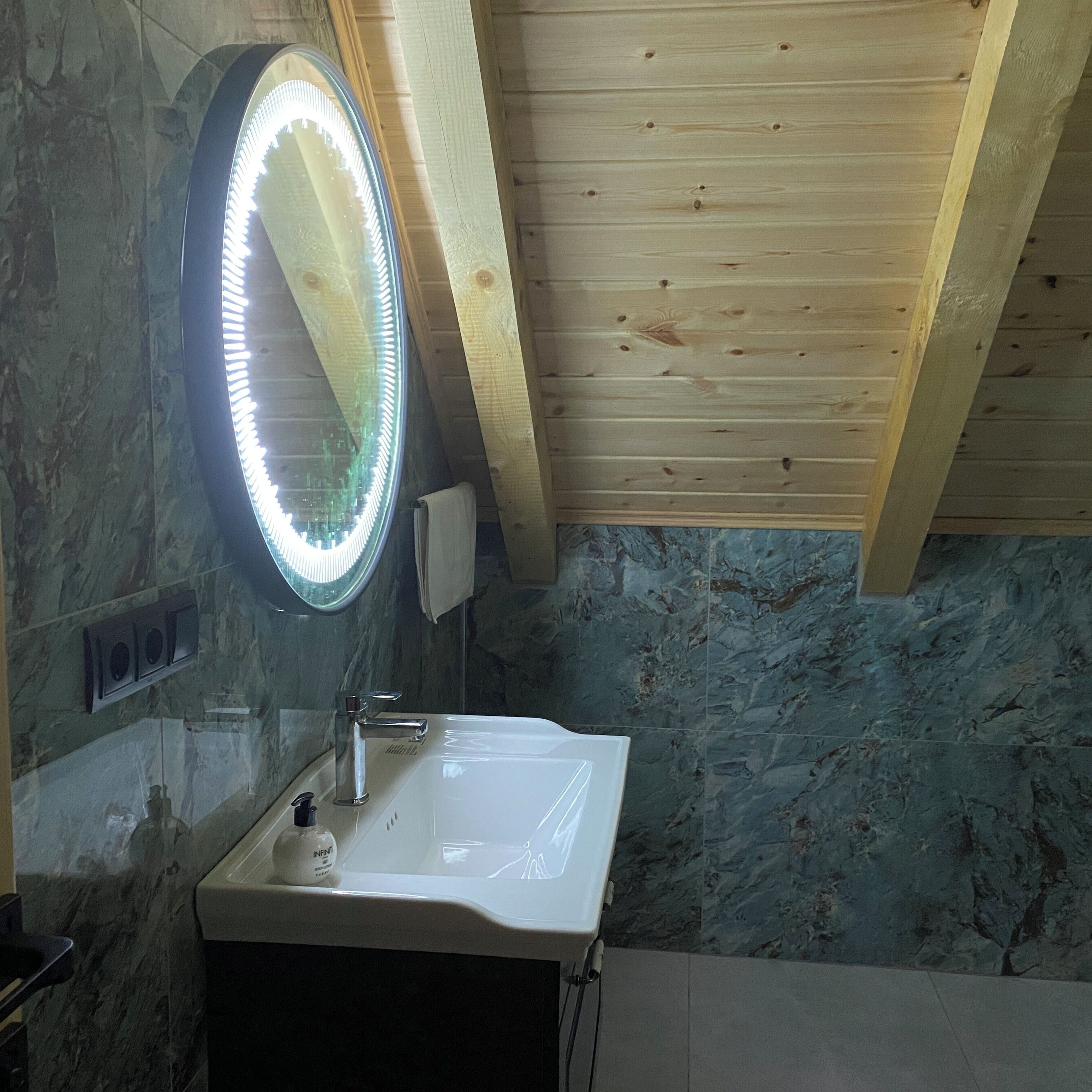 A bathroom with a marble wall, a wooden ceiling, a white sink, and a round mirror with lights.