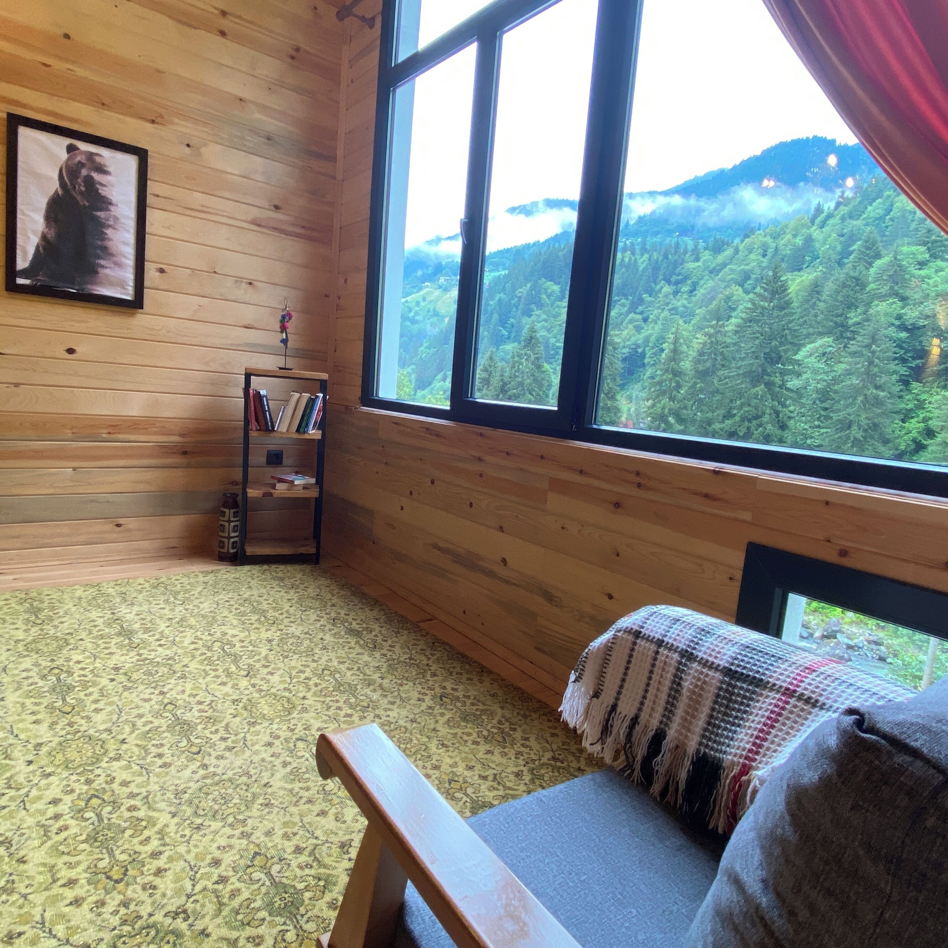 A cozy room with wooden walls, a large window offering a forest view, a bookshelf, and a patterned carpet.