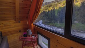 A wooden cabin room with a large window overlooking a river and forested mountains. - Nanu's Ayder Bungalow ve Kahvaltı (Rize)