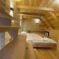 Habitación tipo cabaña de madera con literas, una mesita, un banco y un techo inclinado con tragaluz.
