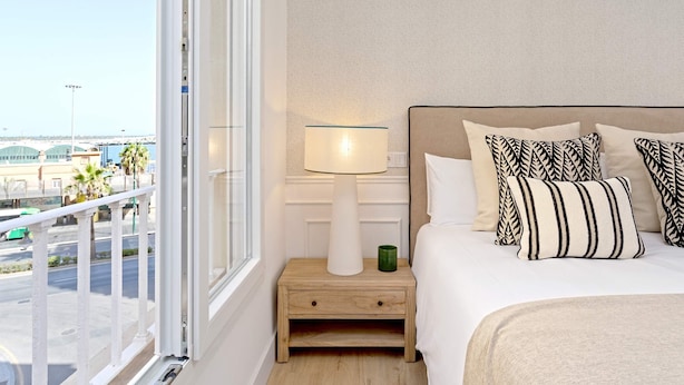 Ett hotellrum med ett stort fönster, ett sängbord med en lampa, en säng med mönstrade kuddar och utsikt över en stadspanorama.