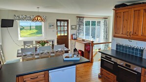 A kitchen with wooden cabinets, a built-in oven, a dining area with a table and chairs, and a window with a view of greenery. - Family Escape | Garden, Parking, Pet friendly (Salcombe)