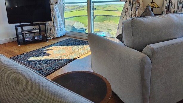 A living room with a large TV, a sofa, a coffee table, and a view of green fields through a large window. - Family Escape | Garden, Parking, Pet friendly (Salcombe)