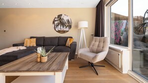 A modern living room with a dark sofa, a wooden coffee table, a beige armchair, and a large window with a view of palm trees and a boat. - 2 bedroom apartment in breukelen (Breukelen)