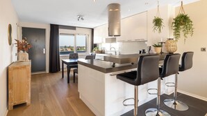 A modern kitchen with a central island, bar stools, hanging plants, and a view through the door. - 2 bedroom apartment in breukelen (Breukelen)