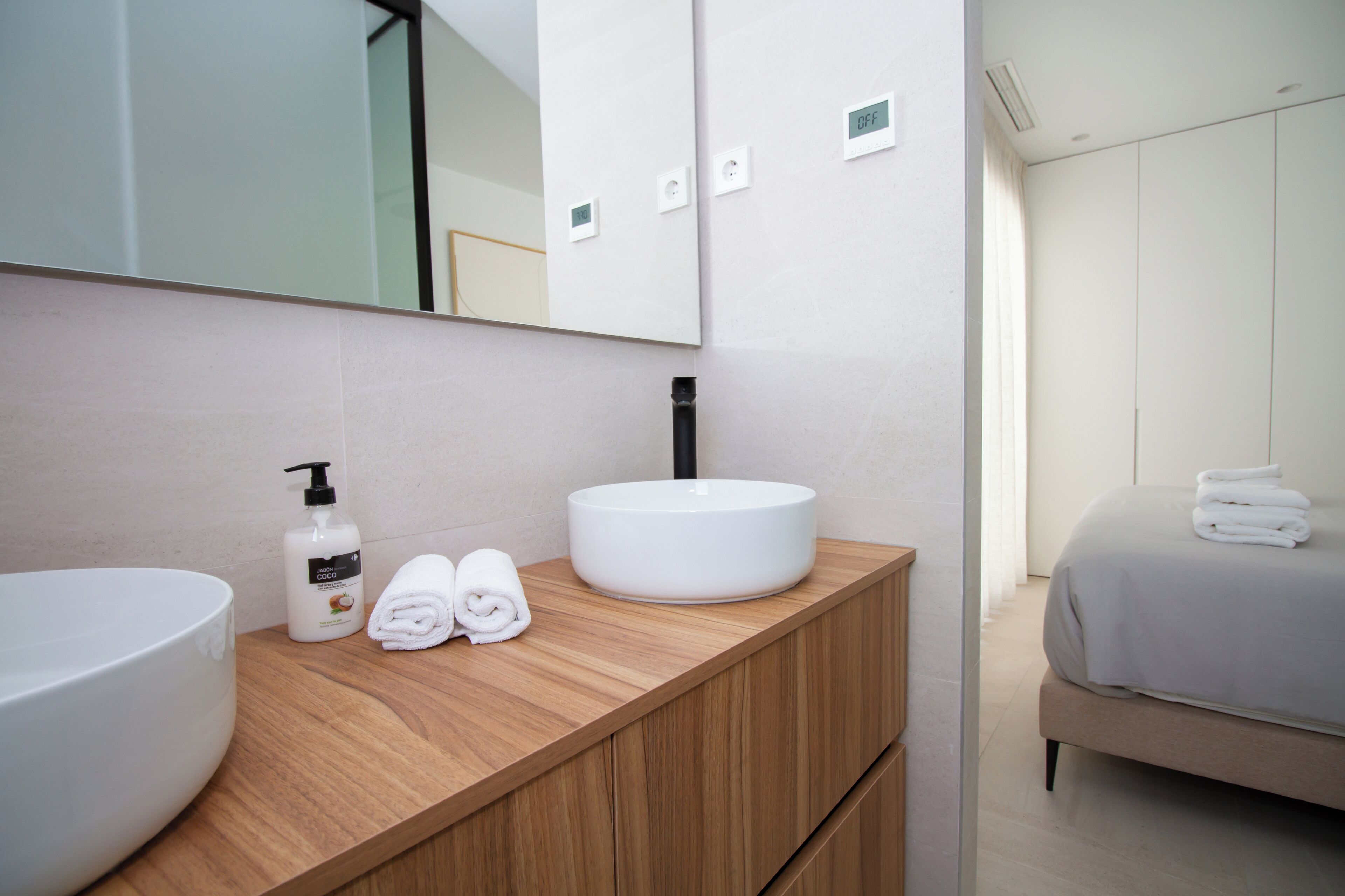 A modern bathroom with a large white sink, a wooden vanity, a mirror, and a bed with a grey bedspread.