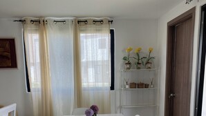 Un comedor con una mesa blanca, sillas y un florero. Hay cortinas, una estantería con plantas y una puerta.