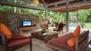 A covered outdoor seating area with a stone wall, a television, and a pool in the background. - 2 BR Tranquil Villa 10 Min Walk to Seminyak Beach (Kuta Utara)