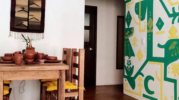 Uma área de refeições com mesa e cadeiras de madeira, parede com padrão geométrico e porta que dá para outro cômodo.