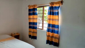 Quarto simples com uma cama, uma pequena mesa de cabeceira de madeira, um relógio de parede e cortinas com padrão listrado.