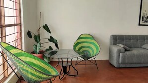 Uma sala de estar com um sofá cinzento, duas poltronas trançadas em verde e amarelo, uma mesa redonda pequena e um vaso com planta.