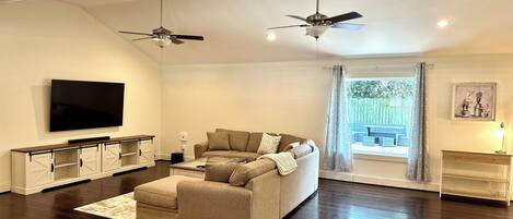 A modern living room with a flat-screen TV, two ceiling fans, a beige sofa, a wooden entertainment unit, and a large window with a view of the outdoors.