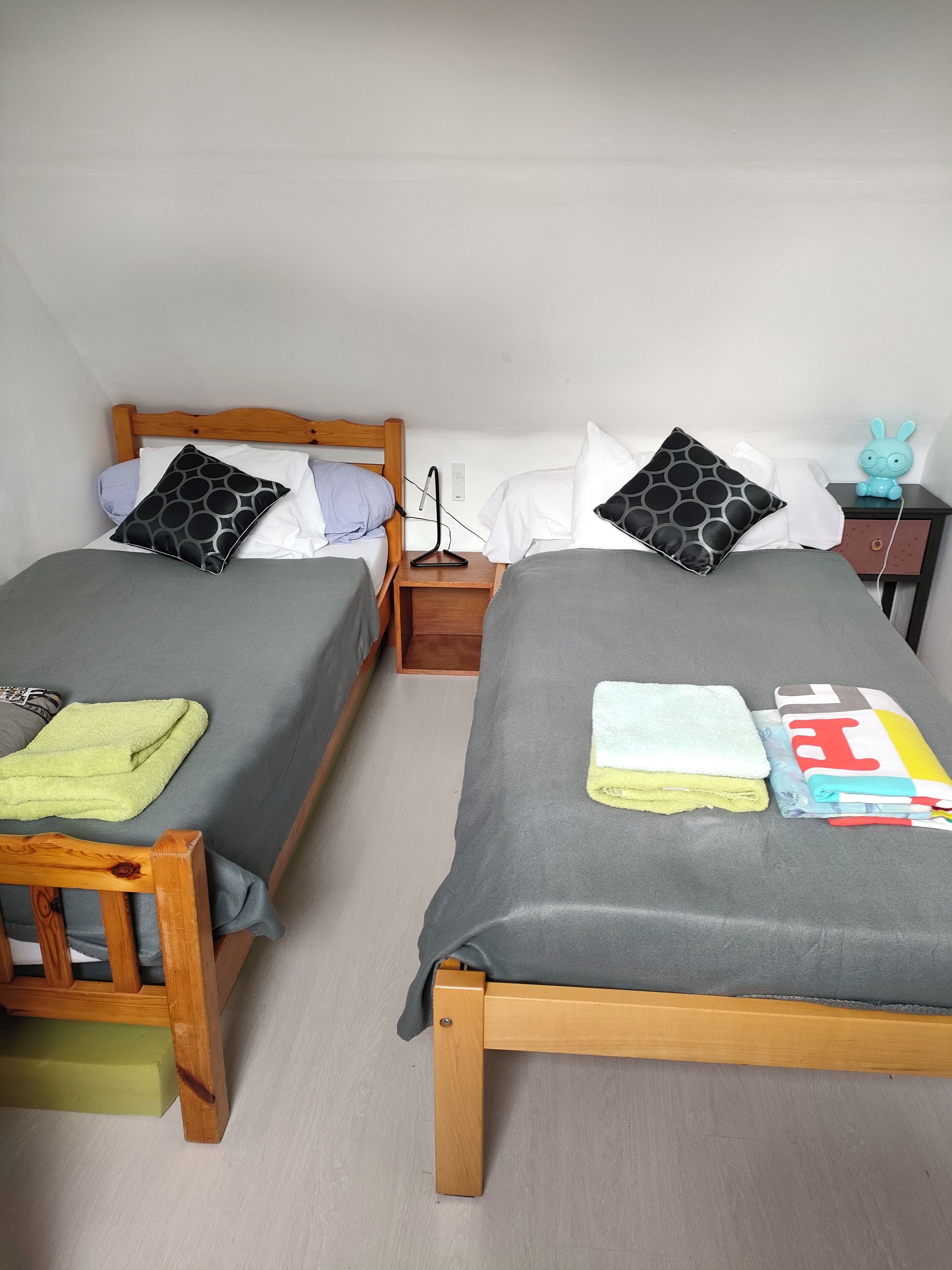 A twin room with two beds, each with a wooden headboard, grey bedding, and patterned pillows. There is a nightstand with a lamp and a small table with a toy figure.