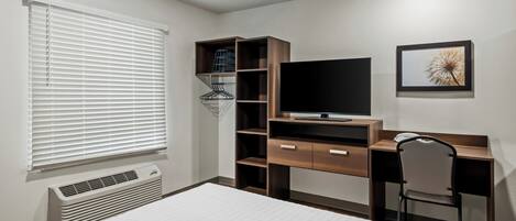 A bedroom with a bed, a desk, a chair, a TV, a heater, and a window with blinds.
