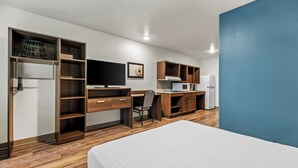 A modern hotel room with a bed, a desk, a chair, a wardrobe, a television, and a kitchenette.