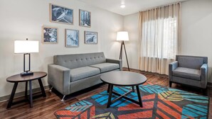 A modern hotel room with a grey sofa, a round coffee table, a side table with a lamp, a patterned rug, and framed pictures on the wall.