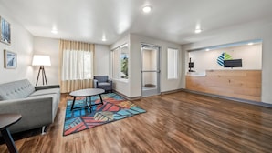 A hotel lobby with a sofa, chairs, a small table, a colorful rug, wooden flooring, and a reception desk.