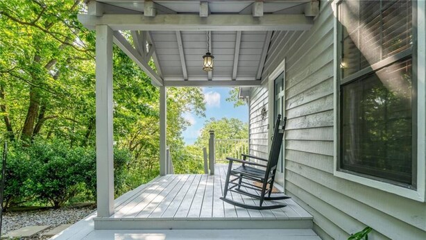 En overdækket veranda med en gyngestol, trapper der fører op til et hus med et vindue, og grøn vegetation i baggrunden.