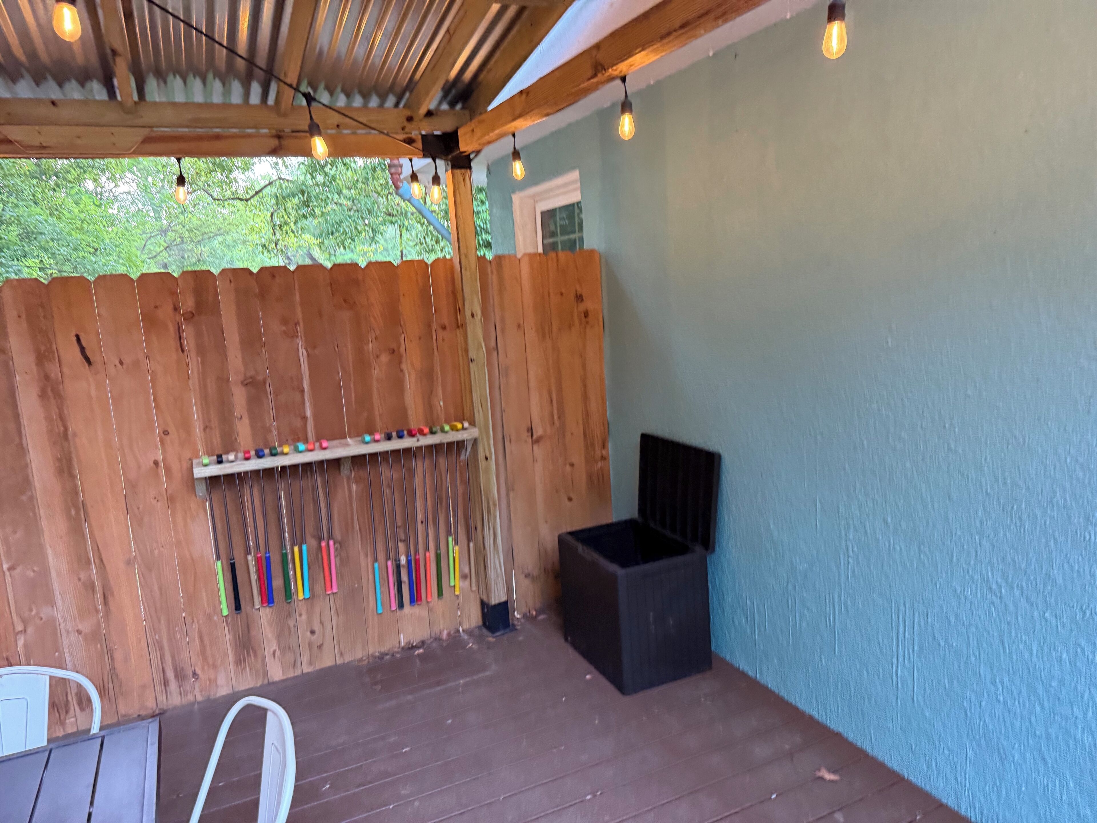 Eine überdachte Terrasse mit Holzlattenzaun, ein Metallregal mit bunten Gegenständen, eine schwarze Mülltonne und hängende Glühbirnen.