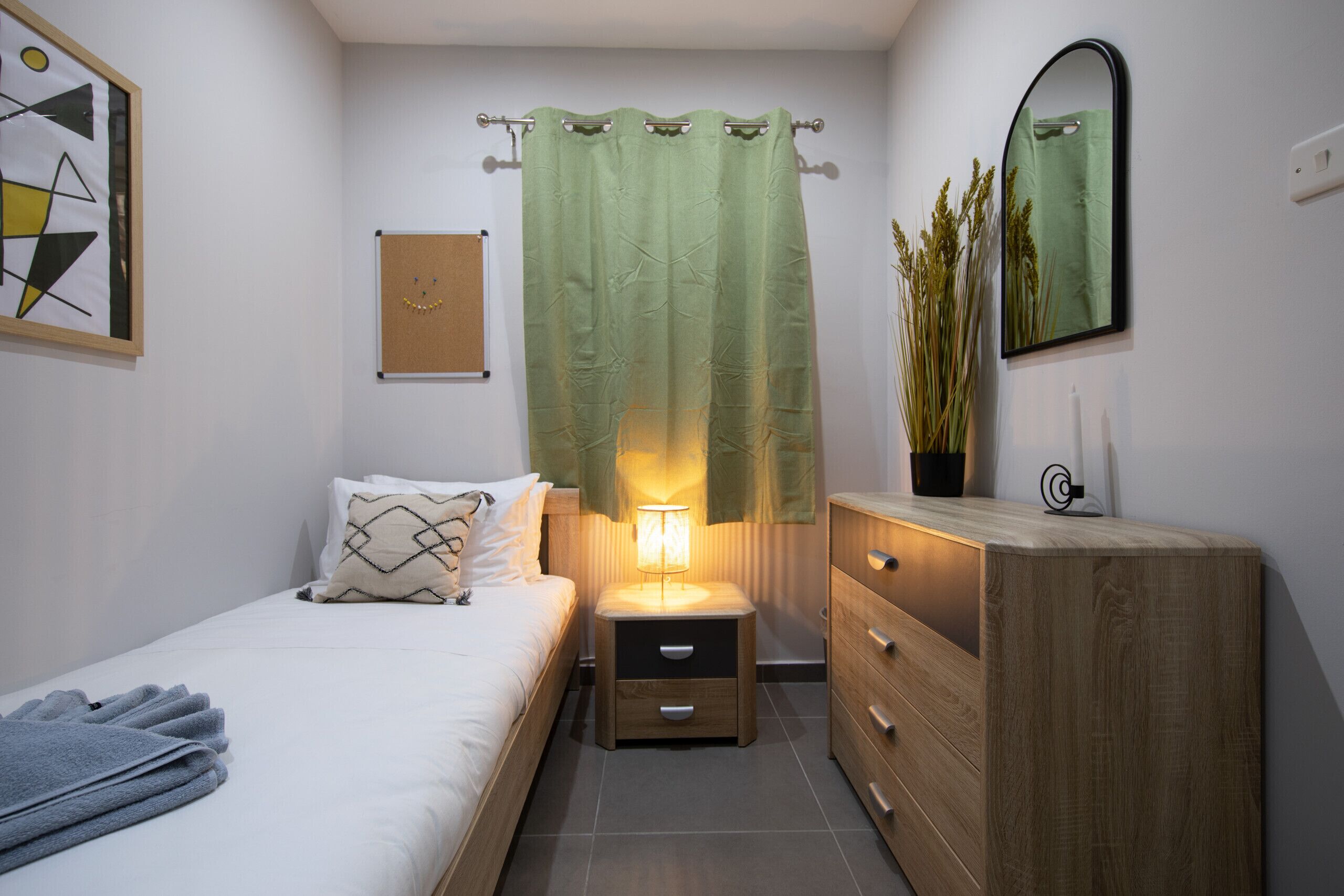 A bedroom with a bed, bedside table with a lamp, a dresser, a mirror, and a green curtain.