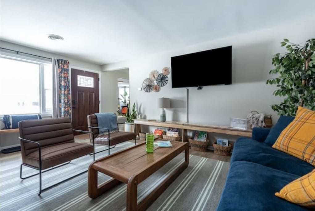 Una sala de estar con sofá, sillón, mesa de centro y televisor de pantalla plana montado en la pared.