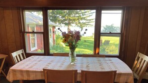 Une table à manger agrémentée d’un centre de table floral, entourée de chaises en bois, placée devant une fenêtre donnant sur un paysage verdoyant et un bâtiment rouge.