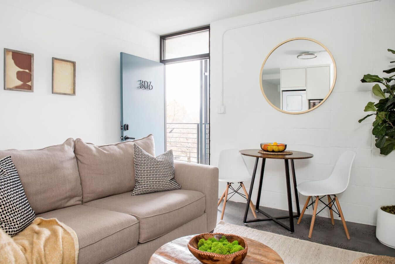 A modern living room with a beige sofa, a round table with a bowl of fruit, a white chair, and a large mirror on the wall.