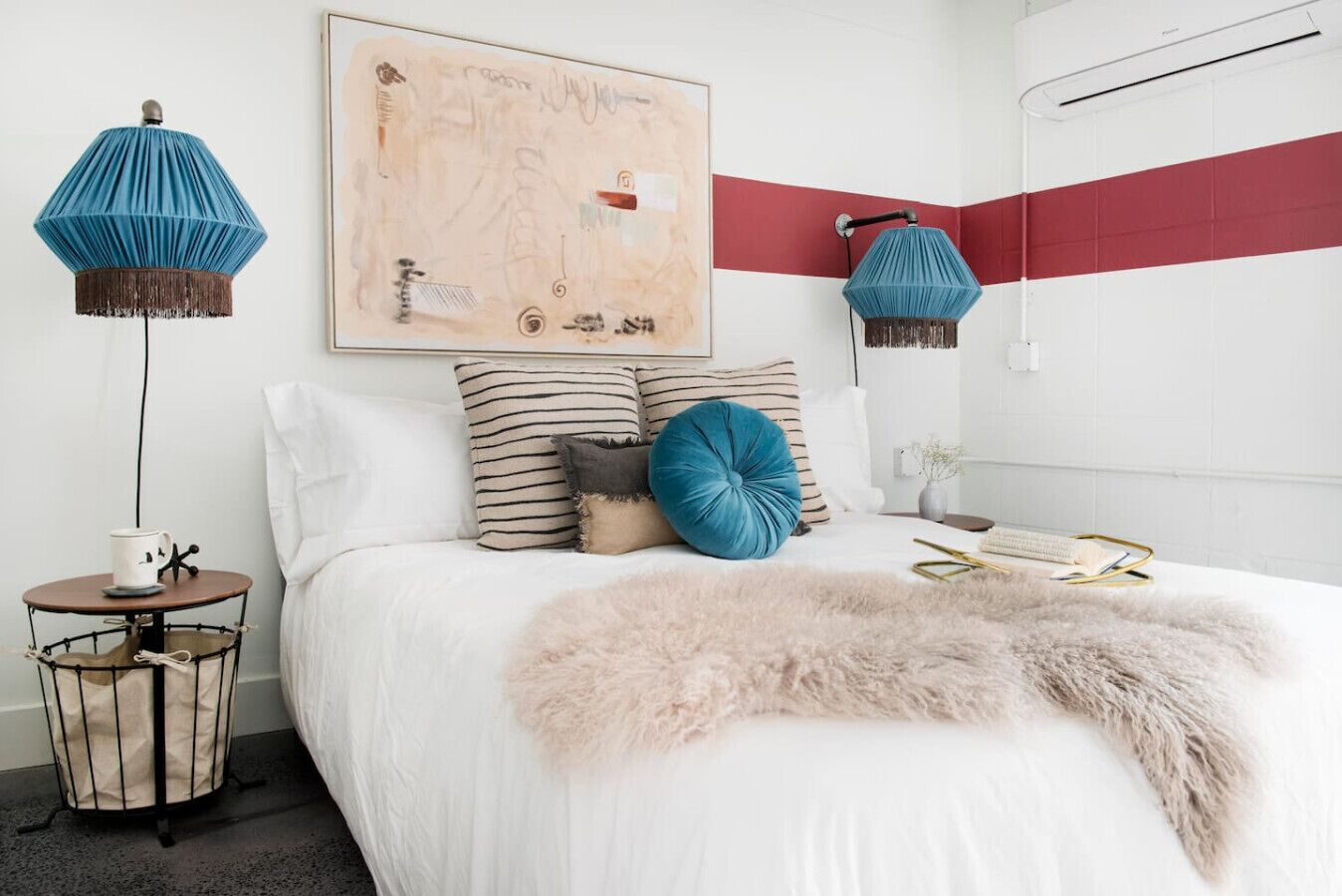 A bedroom with a white bed, striped pillows, a blue accent pillow, a fluffy throw, a round side table with a cup, and two blue lamps.