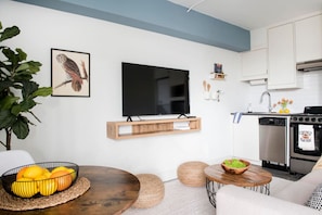 Uma sala moderna com televisão de tela plana, um móvel para TV de madeira, uma mesa de jantar com um cesto de frutas e uma cozinha com armários brancos e fogão.
