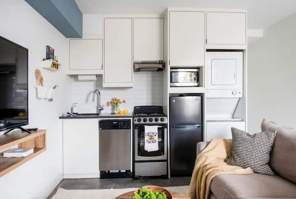 Modern kitchen with flat-screen TV, microwave, oven, and dishwasher, adjacent to a cozy living area with a sofa and throw pillow.