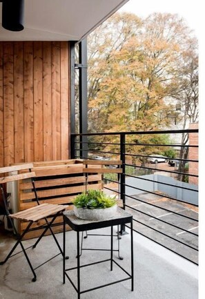 Enjoy the serene view from this balcony with a bench and table, overlooking a tranquil residential street lined with trees.