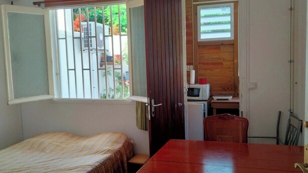 A small, compact room with a bed, a wooden table, a microwave, and a refrigerator. - Studio Lumineux Dans Quartier Calme, Avec Terrasse, 5 mn à Pied du Centre Ville (Basse-Terre)