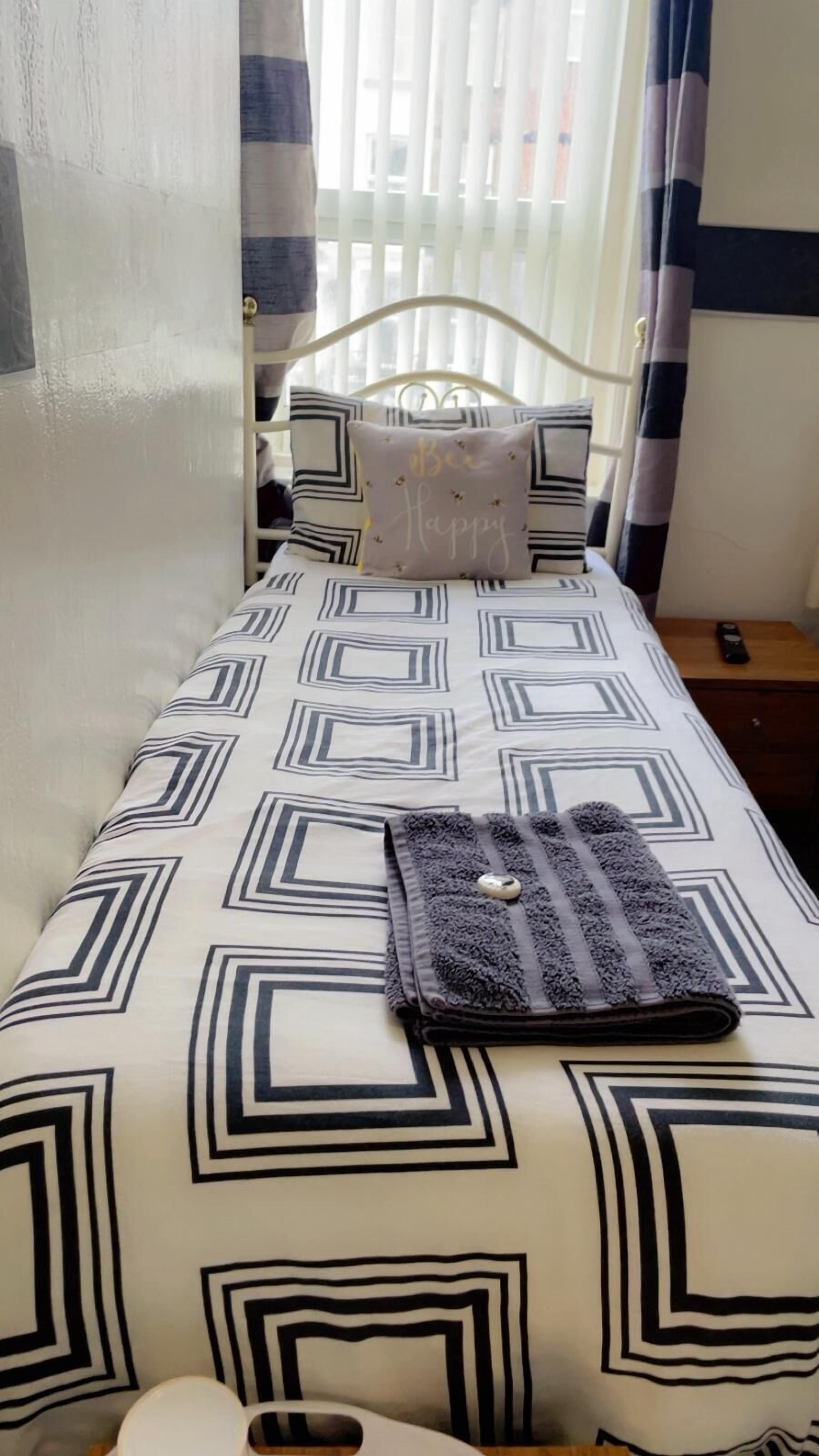 A neatly made bed with a geometric patterned bedspread, a pillow with 'Bee Happy' text, and a folded towel on the bed.