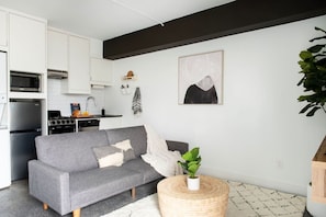 A compact living space with a kitchenette, grey sofa, and a round wooden side table with a plant.