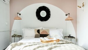 Un dormitorio con cama blanca, dos mesitas de noche, un espejo redondo y una lámpara de pared.
