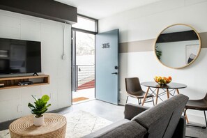 Ein modernes Wohnzimmer mit Fernseher, Sitzgelegenheiten, einem Esstisch mit Obst und einer Tür mit der Aufschrift 305.