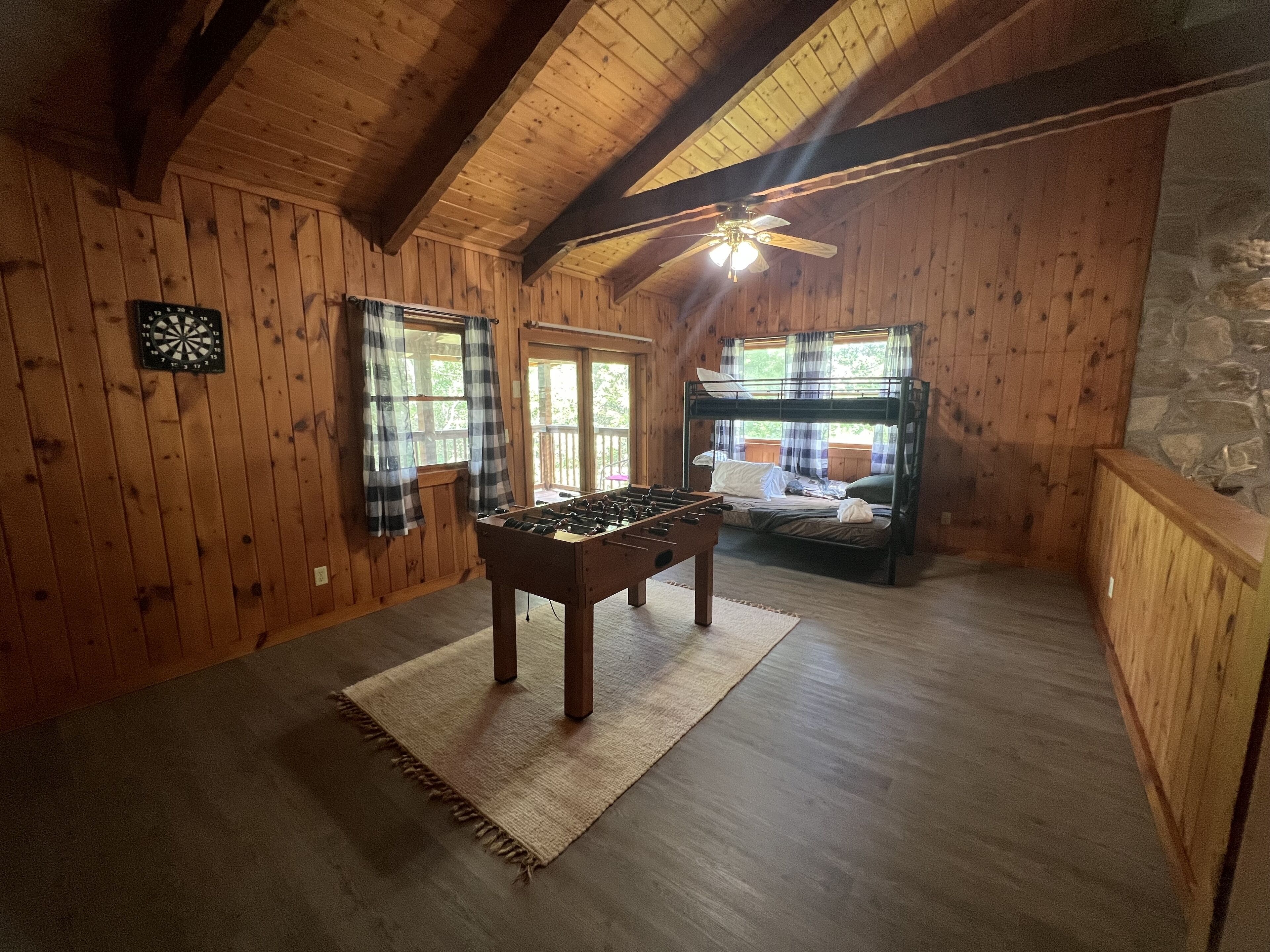 Una habitación acogedora con paredes de madera y techo inclinado, que incluye una mesa de fútbol, una litera y un dardo.