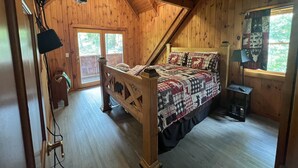 Een gezellige slaapkamer met een houten plafond, een bed met quilt, een nachtkastje, een lamp en een raam met uitzicht op bomen.