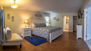 A bedroom with a bed, bedside tables, a chair, a blue rug, and a hallway leading to a bathroom. - Elegant Suite with Jacuzzi (Hancock)
