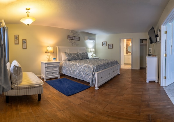 A bedroom with a bed, bedside tables, a chair, a blue rug, and a hallway leading to a bathroom. - Elegant Suite with Jacuzzi (Hancock)