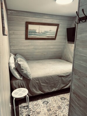 Una habitación pequeña y acogedora con una cama, un taburete, un cuadro en la pared y una pared de madera.